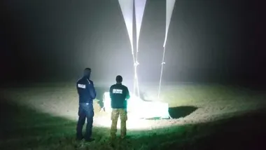 Dwóch funkcjonariuszy stoi nocą na polanie przy rozświetlonym balonie lub sondzie na linach.